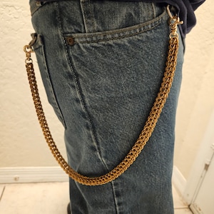 May include: A gold-colored chain wallet leash attached to a pair of blue denim jeans. The chain is made of interwoven links and is secured with a clasp. The jeans are worn by a person.
