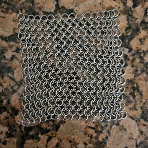 Puede incluir: Un cuadrado de cota de malla plateada brillante. La cota de malla está hecha de pequeños anillos de metal entrelazados, creando una tela flexible en forma de malla. La cota de malla se muestra sobre una superficie moteada marrón y beige.