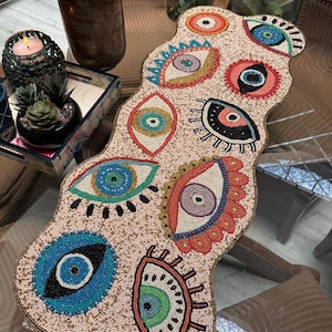 Peut inclure: Chemin de table en perles avec un motif d'œil. Le chemin de table est beige avec des motifs d'yeux perlés colorés en bleu, vert, rouge et jaune. Les bords sont ornés de perles dorées. Cette pièce décorative ajoute une touche unique à toute table.