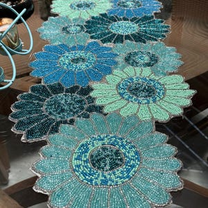 May include: A beaded table runner featuring a floral design. The runner is made of turquoise, teal, and blue beads, arranged in a daisy-like pattern. The petals have a scalloped edge, and the center of each flower is a darker shade of blue.