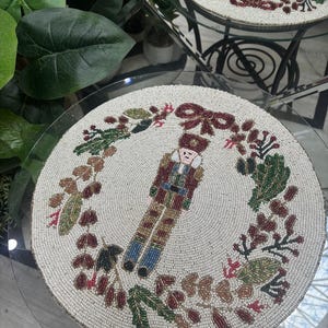 Peut inclure: Nappe ronde perlée avec un soldat casse-noisette au centre, entouré d'une couronne de feuilles et de baies. Le napperon est principalement blanc cassé avec des accents de rouge, de vert, de marron et de bleu. Le design est détaillé et festif.