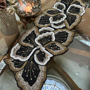 Könnte beinhalten: Ein Tischläufer mit floralem Perlendesign. Der Läufer hat schwarze, weiße und goldene Perlen. Er liegt auf einem Glastisch. Der Tisch ist aus Holz. Der Läufer ist ca. 76 cm lang.