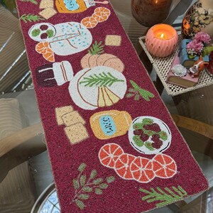 Może przedstawiać: Bordowy bieżnik na stół z koralikami z motywami jedzenia. Bieżnik prezentuje szczegółową pracę z koralików przedstawiającą ser, krakersy, pomarańcze i inne produkty spożywcze. Zielone i pomarańczowe akcenty uzupełniają bordowe tło.