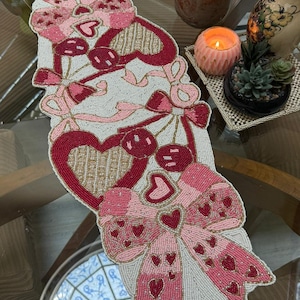 May include: A beaded table runner with a Valentine's Day theme. The runner features a white background with red, pink, and gold heart and bow designs. The hearts and bows are embellished with beads. The runner is placed on a glass table.