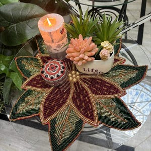 May include: A glass-topped table displays a beaded poinsettia-shaped placemat in burgundy, green, and gold. A lit candle, a small decorative box, and a small white pot with succulents are arranged on the table. The pot has the word "Life" on it.