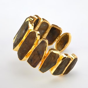 May include: A gold-plated bracelet featuring multiple rectangular, raw-cut stones. The stones have a mottled, earthy appearance with shades of brown and gray. The bracelet's gold setting frames each stone, creating a striking contrast.