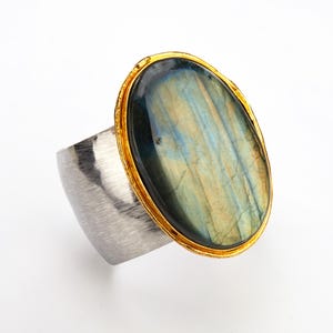 May include: A silver ring with a large oval-shaped labradorite gemstone set in a gold bezel. The ring has a brushed silver finish.