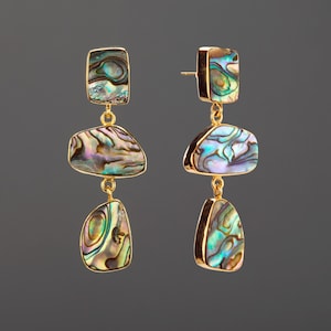 May include: A pair of drop earrings featuring three irregular, polished abalone shell segments set in gold-toned metal. Each earring consists of a rectangular top piece, a wider middle piece, and a smaller bottom piece, all displaying iridescent blue, green, and purple hues.