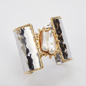 May include: A silver and gold cuff bracelet with a large, white, pearlescent stone set in a gold frame. The bracelet is hammered and has a textured surface.