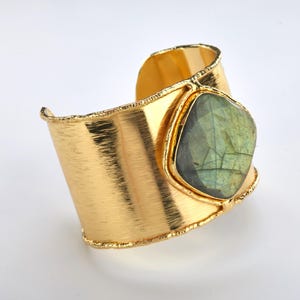 May include: A wide, gold-colored cuff bracelet with a textured finish. The bracelet features a large, square-cut, green and gray gemstone set in a gold bezel. The edges of the bracelet have a rough, textured appearance.
