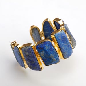 May include: A gold bracelet with a row of rectangular lapis lazuli stones. The stones are a deep blue color and have a slightly irregular shape.