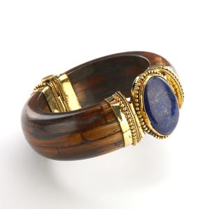 May include: A wooden bangle bracelet with a gold clasp and a large oval blue gemstone. The bracelet is made of dark brown wood with lighter brown streaks. The gemstone is set in a gold bezel.