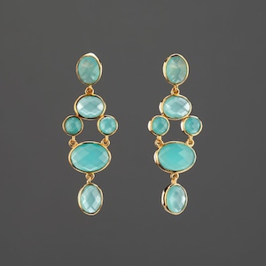 Peut inclure: Une paire de boucles d'oreilles pendantes dorées avec plusieurs pierres précieuses ovales et rondes bleu clair. Les boucles d'oreilles ont un design en cascade, avec des pierres précieuses de tailles et de formes variées. Le fond est gris foncé uni.
