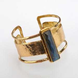 Puede incluir: Pulsera de brazalete de oro con una piedra rectangular azul engastada en el centro.