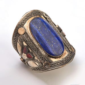 Op de afbeelding: Een zilveren armband met een ovale lapis lazuli-steen in het midden. De armband is versierd met ingewikkelde zilveren filigraan en rode en gouden accenten.