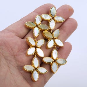 May include: A pair of gold-toned drop earrings featuring a textured metal finish and multiple marquise-shaped mother-of-pearl inlays arranged in a floral or leaf-like cluster pattern, creating a layered and dangling design with iridescent white accents.