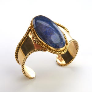 Op de afbeelding: Goudkleurige manchetarmband met een grote, ovale lapis lazuli steen. De steen is gezet in een gouden bezel en de armband heeft een touwachtig ontwerp langs de randen. De manchet is aan de onderkant open.
