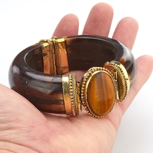 Puede incluir: Un brazalete de madera marrón oscuro con detalles metálicos dorados y una gran piedra ojo de tigre ovalada. El brazalete se sostiene en una mano sobre un fondo blanco. El brazalete mide aproximadamente 6,35 cm de ancho.