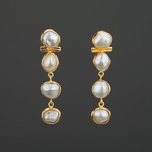 Pode incluir: Um par de brincos pendurados dourados com três pérolas brancas de formato irregular. As pérolas são suspensas por uma barra dourada e conectadas por delicadas correntes douradas. Design clássico e elegante.