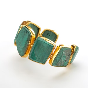 May include: A cuff bracelet featuring multiple irregular, rectangular-shaped green malachite stones set in polished gold-tone metal frames. The stones display natural dark green banding and variations in texture, arranged in a continuous open-ended band.