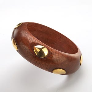 May include: A dark brown wooden bangle bracelet with gold-colored metal accents. The bracelet is a thick, rounded band with several evenly spaced, circular and teardrop-shaped gold embellishments.