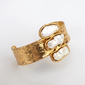 Peut inclure: Un bracelet manchette doré avec une surface texturée. Il présente trois perles d'eau douce blanches de forme irrégulière, serties dans des cadres dorés. Le design est organique et élégant, idéal pour les occasions spéciales.