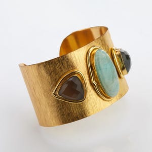 May include: A gold-toned cuff bracelet with a textured surface. The bracelet features three gemstones: a brown triangular stone, a light blue oval stone, and a dark green, faceted stone. The gemstones are set in gold bezels.