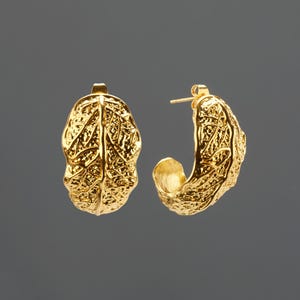 May include: A pair of gold-colored hoop earrings. Each earring has a textured, leaf-like design. The earrings are set against a gray background, highlighting their intricate details and metallic sheen. The earrings are a jewelry item.