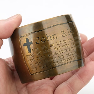 May include: A wide, cylindrical bronze-toned cuff bracelet featuring an engraved cross and the text John 3:16, For God so loved the world that he gave his only begotten Son, that whosoever believeth in him should not perish, but have everlasting life.