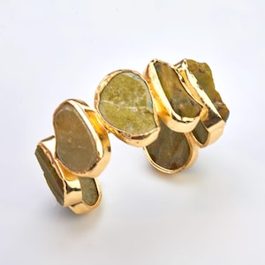 May include: A gold-plated bracelet featuring irregularly shaped, olive-green gemstones. The stones are set in a gold bezel setting, creating a striking contrast. The bracelet is designed with a flexible, articulated construction, allowing it to conform to the wrist.