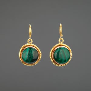 May include: A pair of gold-tone earrings with a round, green malachite stone. The earrings have a simple design with a gold-colored frame around the stone and a hook closure. The malachite has a dark green color with lighter green swirls.