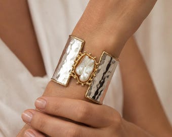 May include: A wide cuff bracelet featuring a hammered silver-tone metal band with gold-tone edges. The center displays an elongated, irregular white pearl set within a gold-tone frame adorned with small, rounded metal beads along its perimeter.