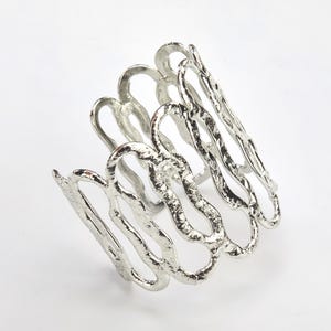May include: A silver bracelet with a wavy, organic design. The bracelet is made of metal and has a textured surface.