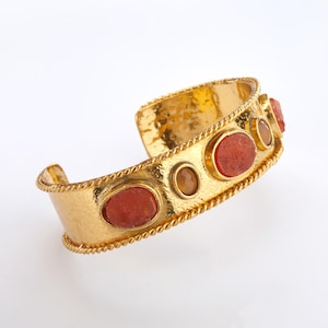 May include: Gold-coloured cuff bracelet with a hammered texture and rope-like trim. The bracelet features oval and round reddish-orange and tan-coloured stones. The open design allows for easy wear and adds a touch of elegance.