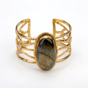 May include: Gold-toned cuff bracelet featuring an oval, iridescent gemstone. The bracelet has an open design with intricate, layered metalwork. The gemstone displays shades of blue, green, and brown, set in a gold bezel.
