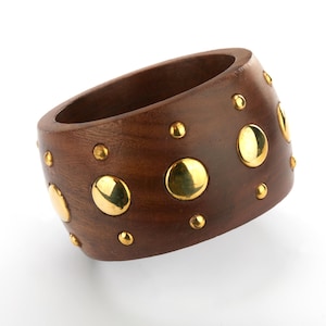 May include: A dark brown wooden bangle bracelet with gold-colored metal studs. The bracelet is thick and has a smooth, polished finish. The gold studs are various sizes and are evenly spaced around the bracelet.