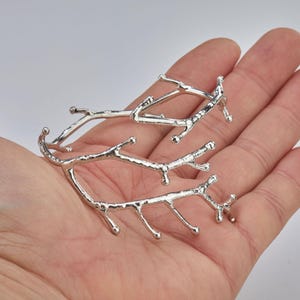 Könnte beinhalten: Silberfarbene, astförmige Schmuckstücke. Der Schmuck hat eine strukturierte Oberfläche und ein natürliches, organisches Design. Die Stücke werden in einer Hand gehalten, um ihre Größe und Details zu zeigen. Die Gesamtästhetik ist minimalistisch und von der Natur inspiriert.