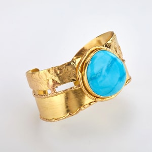 May include: A gold-coloured cuff bracelet featuring a large, oval turquoise stone. The bracelet has a textured, hammered finish on the top and a smooth, polished finish on the band. The turquoise stone is set in a gold bezel.