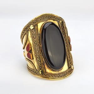 May include: A wide, gold-toned cuff bracelet with an oval, black stone centerpiece. The bracelet features intricate filigree and small red accent stones. The design has a vintage or ethnic aesthetic.
