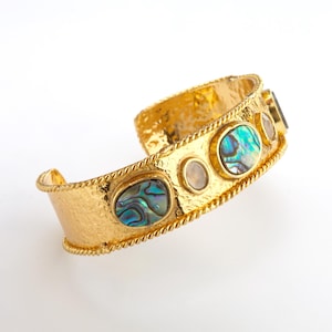 Op de afbeelding: Een goudkleurige manchetarmband met een gehamerde textuur en een touwachtige rand. De armband heeft verschillende ovale en ronde iriserende stenen in tinten blauw, groen en wit. Een stijlvol sieraad.