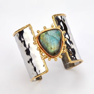 May include: A wide cuff bracelet featuring a hammered silver-tone metal band with gold-tone edges. Centered on the band is a large, triangular labradorite gemstone set in a gold-tone frame adorned with small decorative metal spheres.