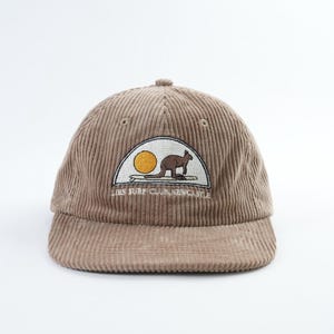 May include: A brown corduroy baseball cap with a curved brim. The cap features an embroidered patch with a kangaroo silhouette, a sun, and the text "LIES SURF CLUB, NEWCASTLE". The cap has a structured crown and a button on top.