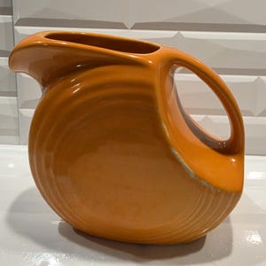 May include: Orange ceramic pitcher with a curved handle and spout. The pitcher has a glossy finish and a rounded body with a ridged texture. The interior is a darker shade of orange. A classic kitchenware item.