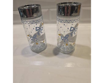 May include: Two clear glass shakers with shiny silver-tone metal lids. Each shaker features a repeating pattern of gray geese with blue bows, yellow beaks, and small blue raindrops. The top and bottom borders have a blue geometric zigzag design.