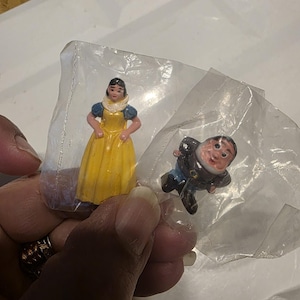 May include: Two miniature figurines, one of a woman in a yellow dress with blue sleeves and a white collar, and another of a man in a black suit and blue pants. Both are in clear plastic packaging.