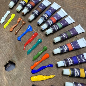 K&ouml;nnte beinhalten: Ein Set mit 10 Acrylfarbtuben in verschiedenen Farben, darunter Wei&szlig;, Gelb, Braun, Rot, Gr&uuml;n, Blau, Orange, Schwarz und zwei Gr&uuml;nt&ouml;ne. Die Tuben sind in einer Reihe auf einer Holzoberfl&auml;che angeordnet, wobei die Farbe aus jeder Tube herausgedr&uuml;ckt wurde, um die Farbe zu zeigen.