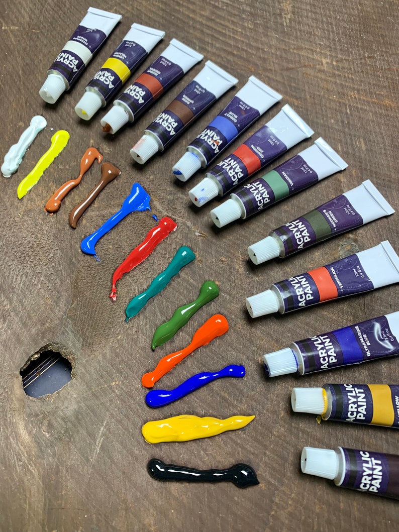 K&ouml;nnte beinhalten: Ein Set mit 10 Acrylfarbtuben in verschiedenen Farben, darunter Wei&szlig;, Gelb, Braun, Rot, Gr&uuml;n, Blau, Orange, Schwarz und zwei Gr&uuml;nt&ouml;ne. Die Tuben sind in einer Reihe auf einer Holzoberfl&auml;che angeordnet, wobei die Farbe aus jeder Tube herausgedr&uuml;ckt wurde, um die Farbe zu zeigen.