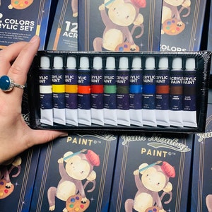 K&ouml;nnte beinhalten: Ein Set mit 12 Acrylfarben in Tuben, mit einer Affenillustration auf der Verpackung. Die Farbtuben sind mit den Farbnamen beschriftet und haben ein schwarzes und goldenes Design. Die Verpackung tr&auml;gt den Schriftzug "June Monkey Paint" und "12 Farben Acryl Set".