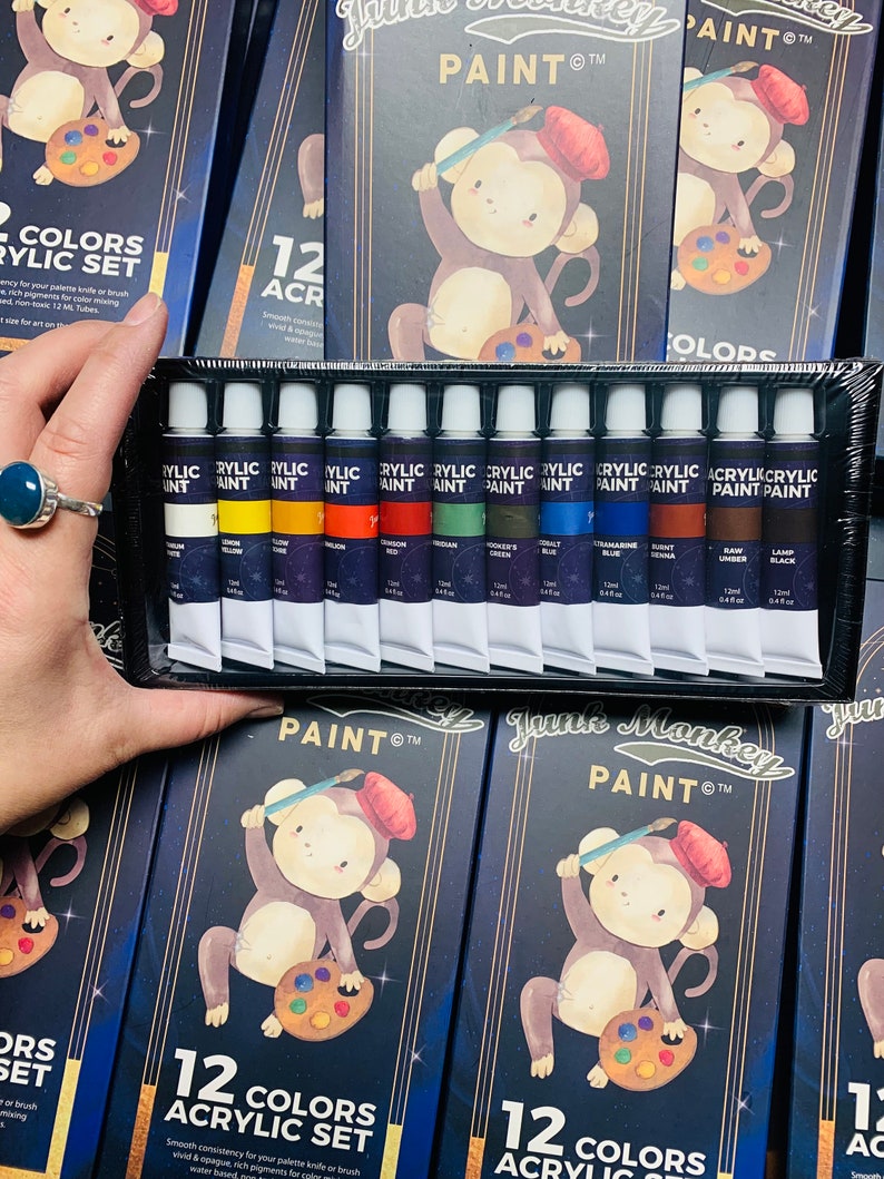 K&ouml;nnte beinhalten: Ein Set mit 12 Acrylfarben in Tuben, mit einer Affenillustration auf der Verpackung. Die Farbtuben sind mit den Farbnamen beschriftet und haben ein schwarzes und goldenes Design. Die Verpackung tr&auml;gt den Schriftzug "June Monkey Paint" und "12 Farben Acryl Set".
