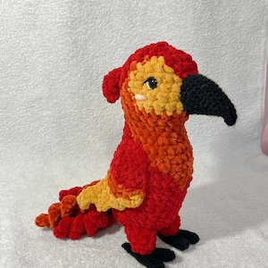 May include: A handmade crochet parrot in vibrant red, orange, and yellow. The plush bird has a black beak and feet, with a detailed tail and head. The soft, textured yarn creates a cuddly appearance, perfect for a child's toy or home decor.